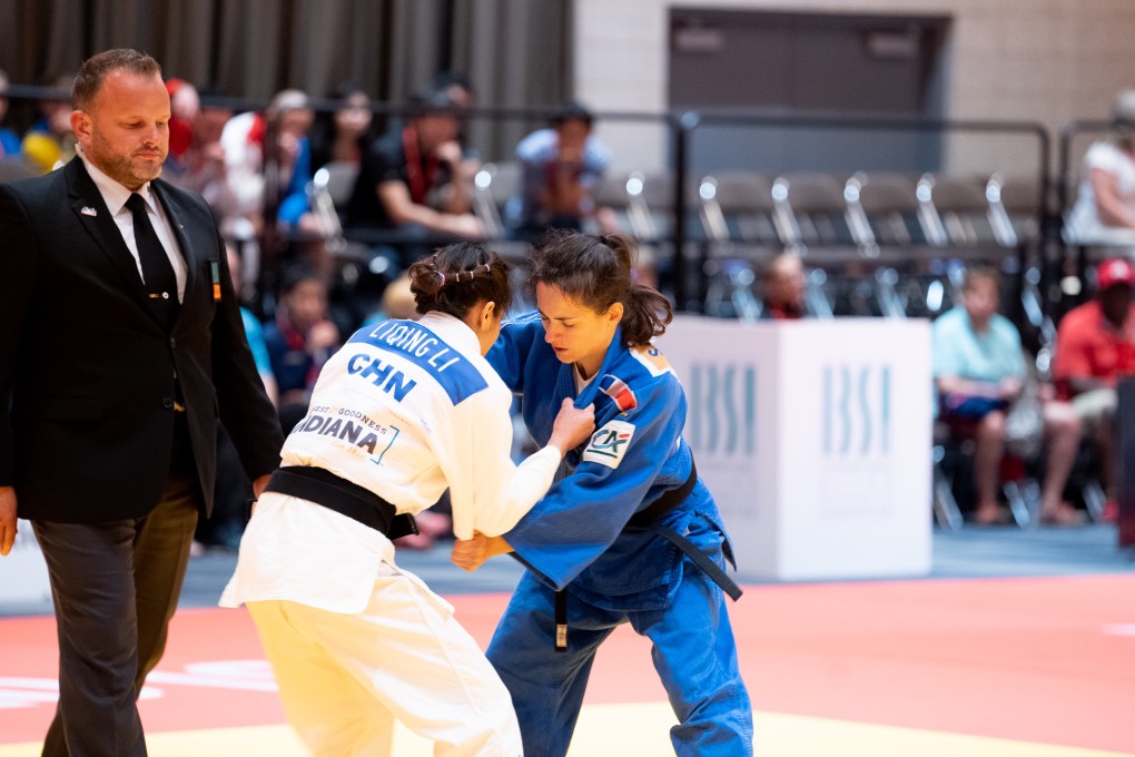 Two female judoka are pictured in a grip on the mat with the referee watching on