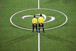 Two referees stand on the centre circle of the blind football pitch at Rio 2016 with the Paralympic symbol in the circle