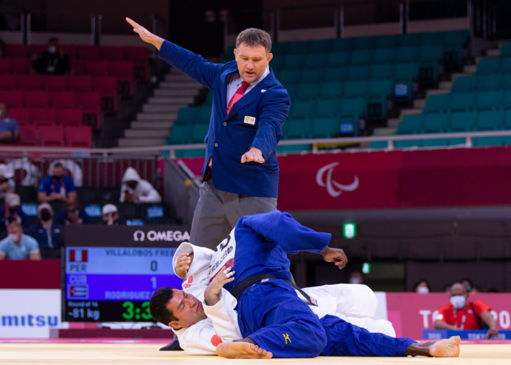 Two judoka are pictured on the mat in a hold. The referee is signalling an ippon with his hand