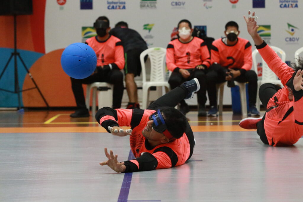 A male goalball player from Mexico stretches across the court to reach out for a save, watched by his teammates on the bench